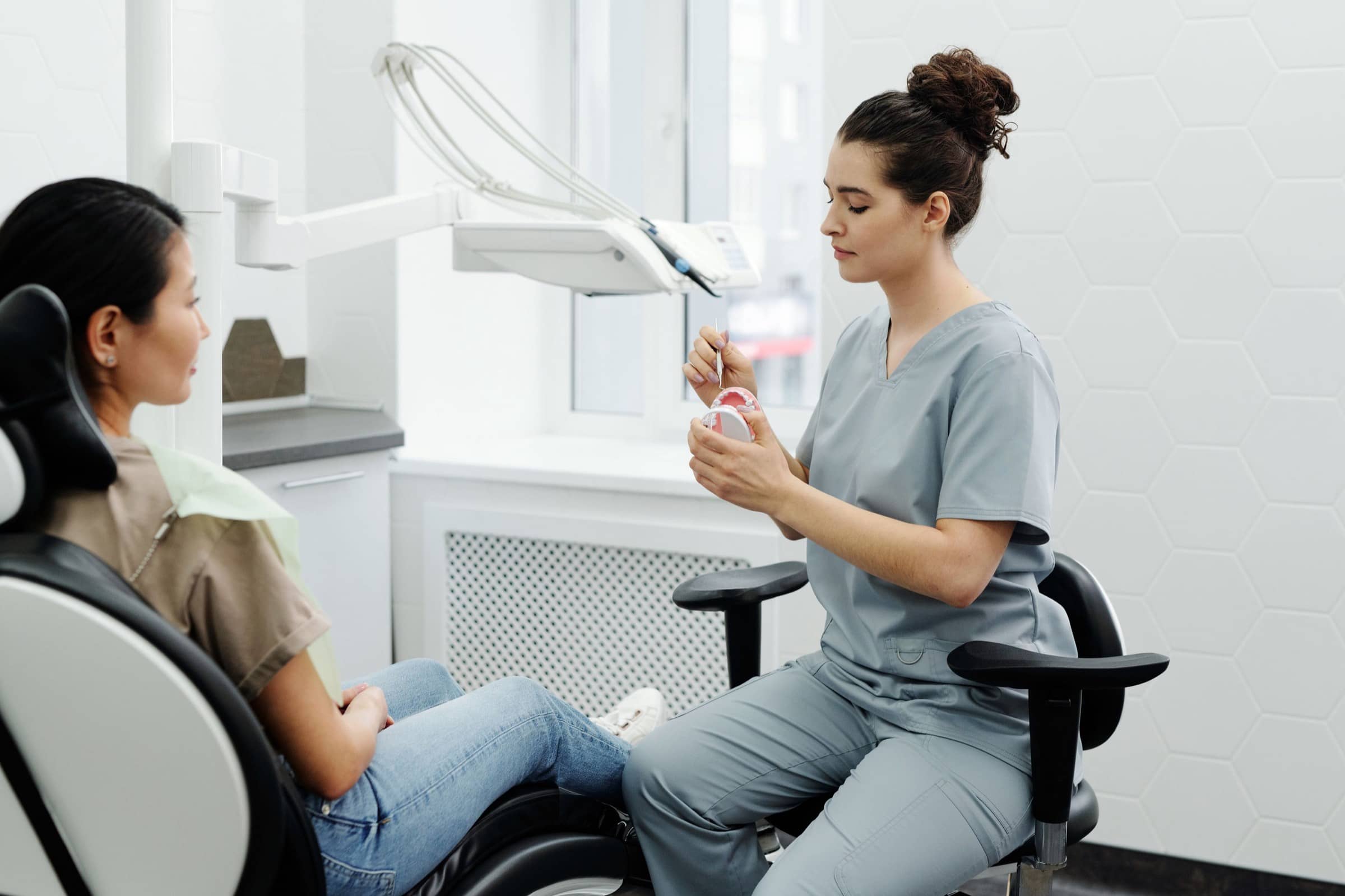 A dentist in grey scrubs talks a seated patient through a dental model.