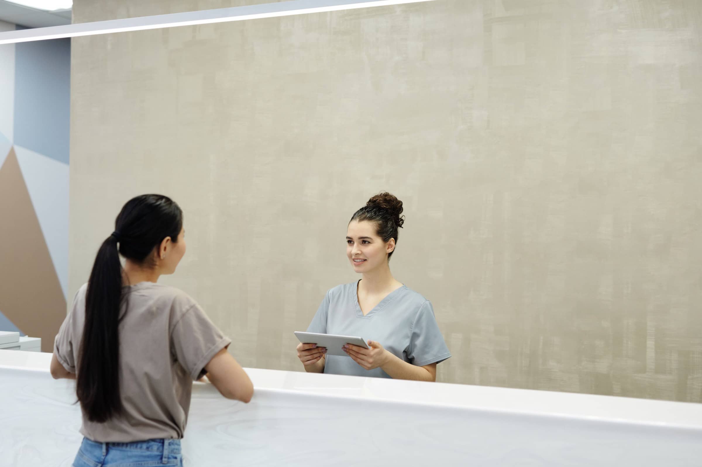 A receptionist in scrubs greets a patient at a clinic front desk.