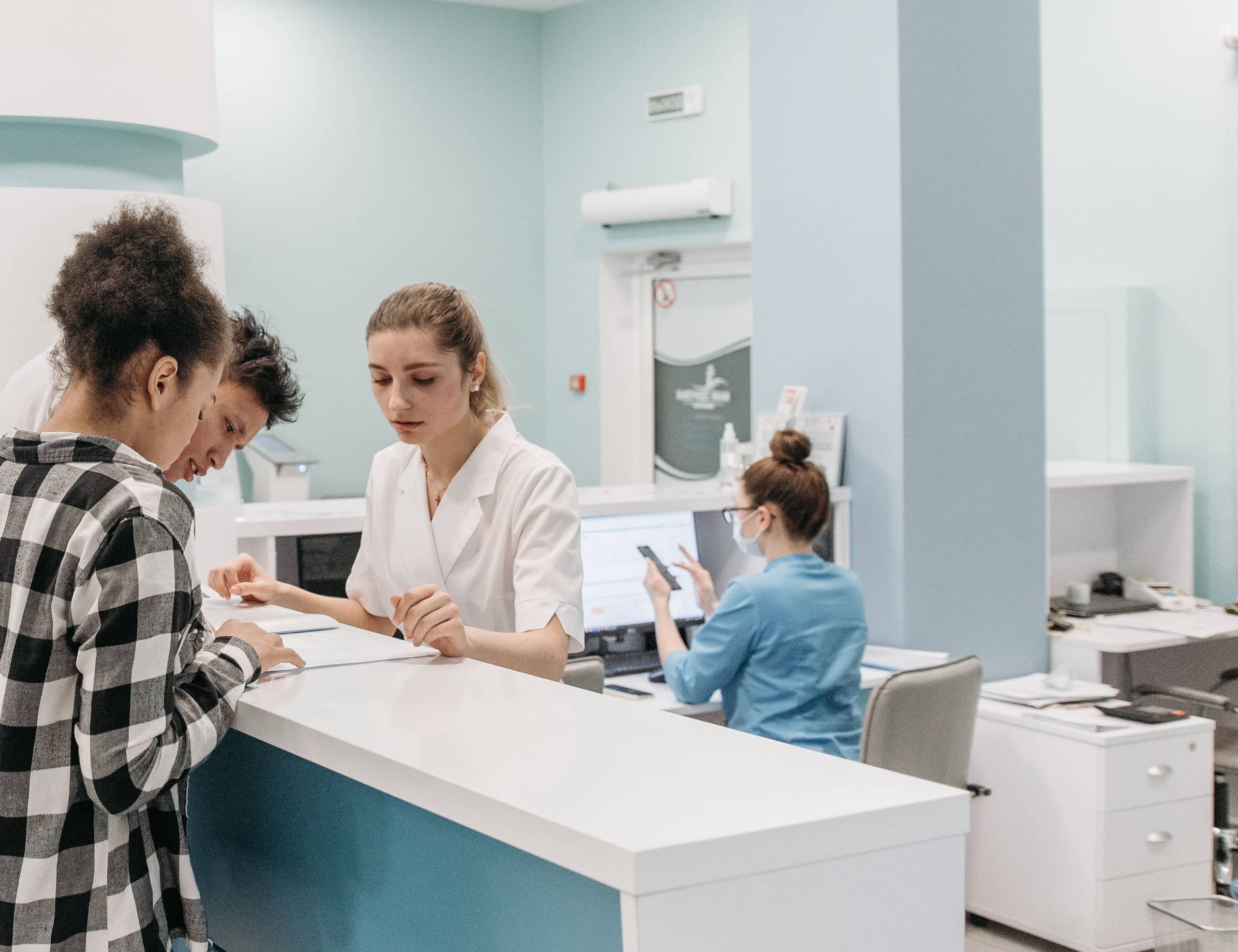Two patients at a clinic reception counter with a receptionist, while a second staff member behind the desk holds a phone.