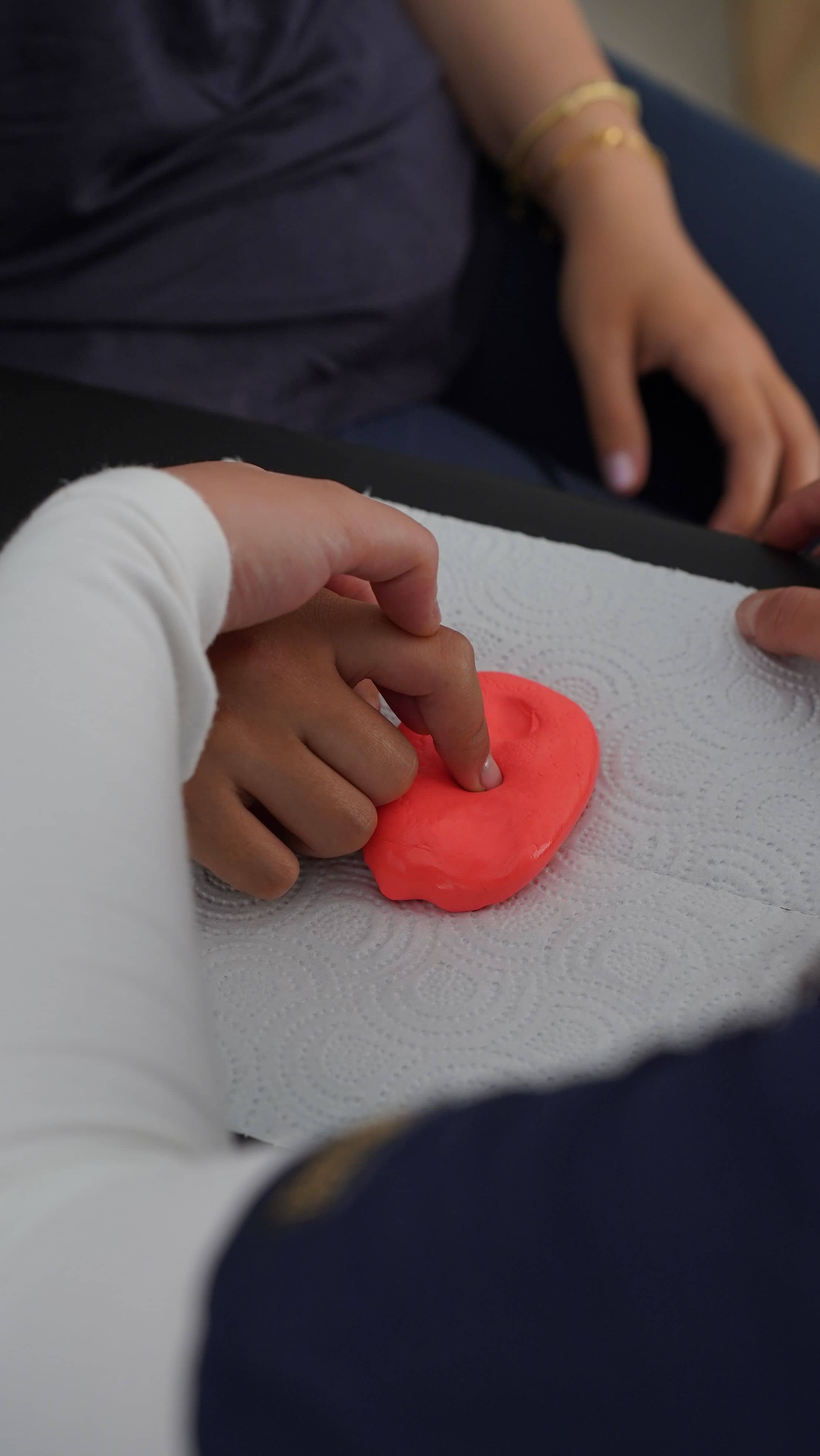 A hand presses a finger into a lump of therapy putty on a table, with a second person’s hands resting nearby.