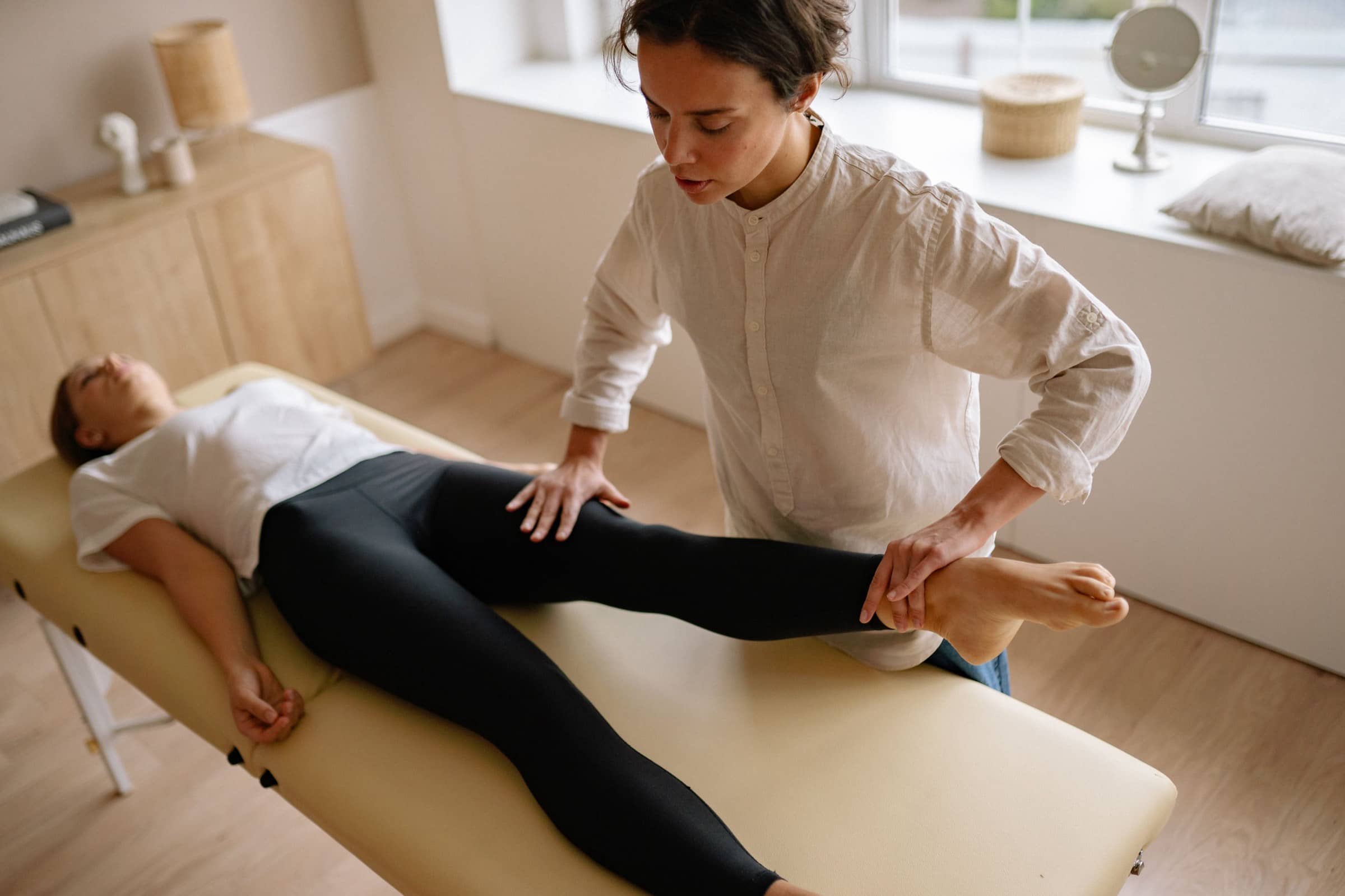 A physiotherapist in a linen shirt holds a patient’s lower leg on a treatment table in a sunny room.
