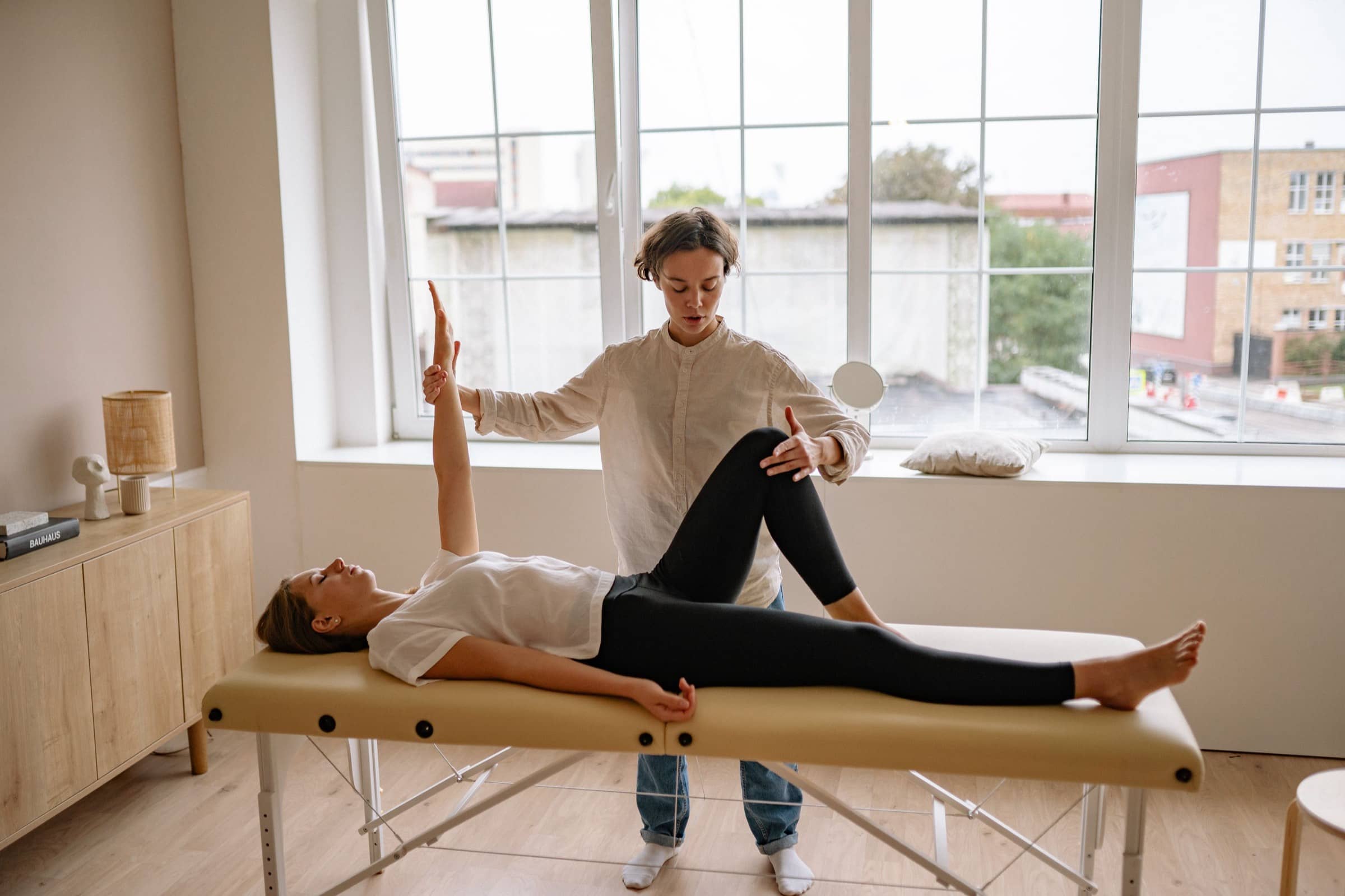 A physiotherapist guides a patient’s arm and knee on a treatment table by tall windows