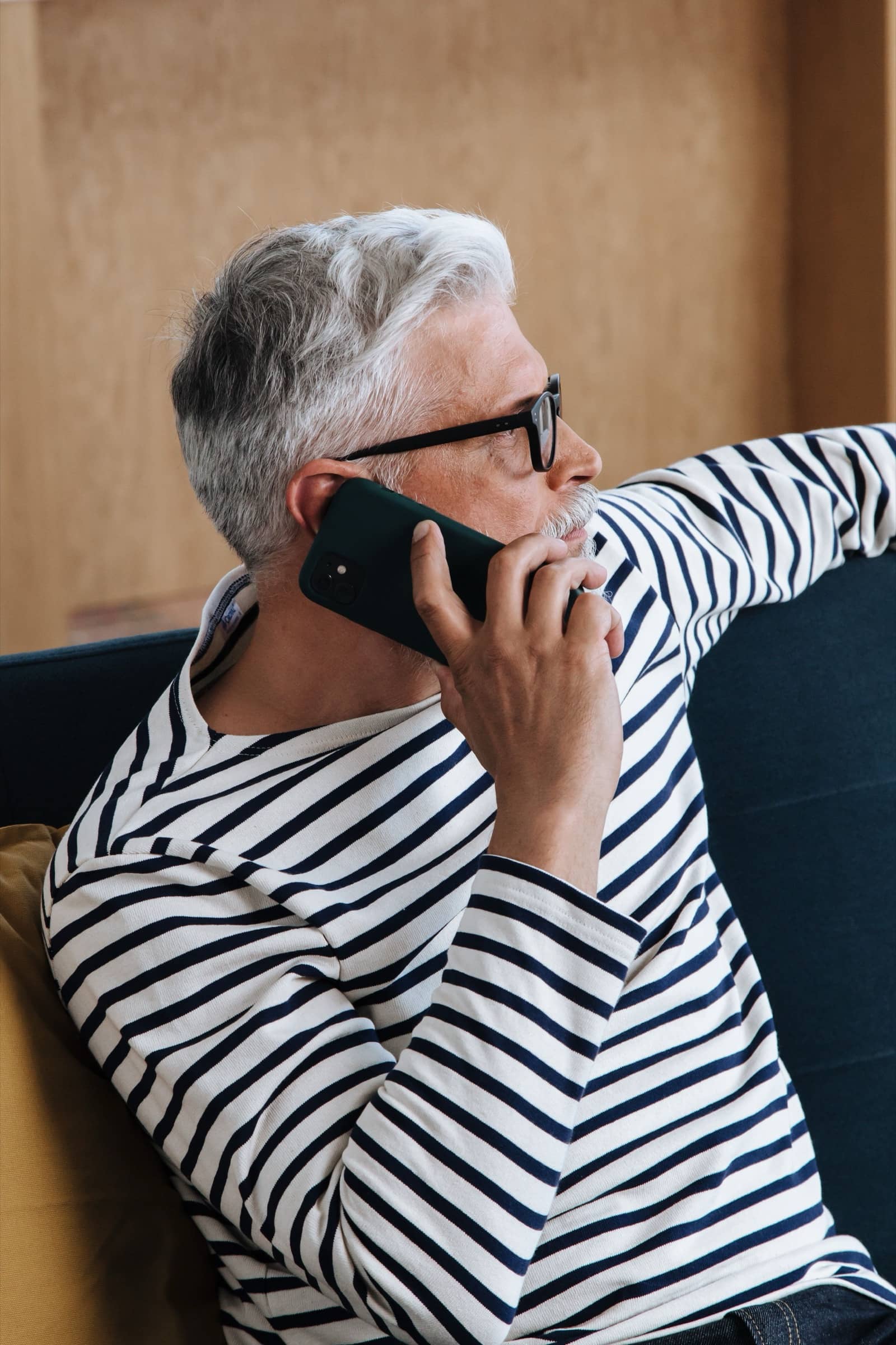 A grey-haired man in a striped top on the phone on his sofa